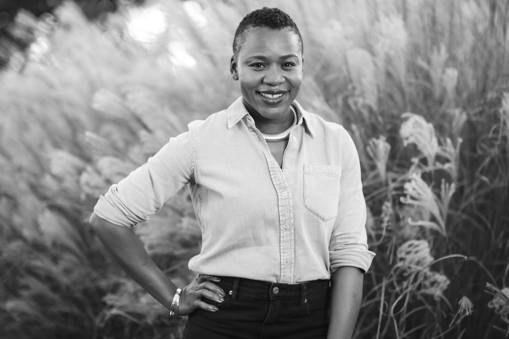 Black and white portrait of a person smiling, standing outdoors with tall grass in the background. They are wearing a light-colored shirt and dark pants, with one hand on their hip.