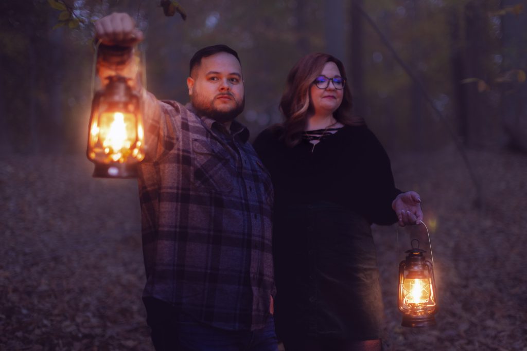 Two people stand in a dimly lit forest at dusk, holding lanterns. The man wears a plaid shirt and the woman, in glasses and a dark sweater, gazes ahead. The scene has an eerie, almost spooky quality to it, like a live-action portrait of mystery.