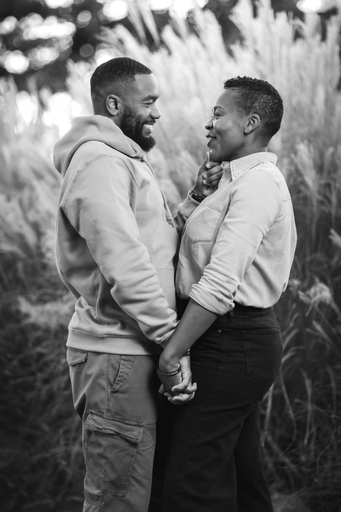 A couple stands outdoors in front of tall grass, holding hands and smiling at each other. In this charming portrait, the man wears a hoodie and cargo pants, while the woman sports a button-up shirt and jeans.
