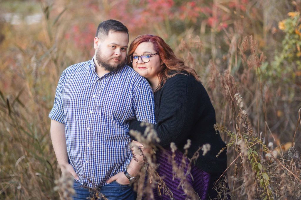 A bearded man in a checkered shirt stands next to a woman with red hair and glasses in a black sweater, leaning on his shoulder, surrounded by tall grass and autumn-colored foliage, creating moody portraits that capture the essence of a spooky fall day.