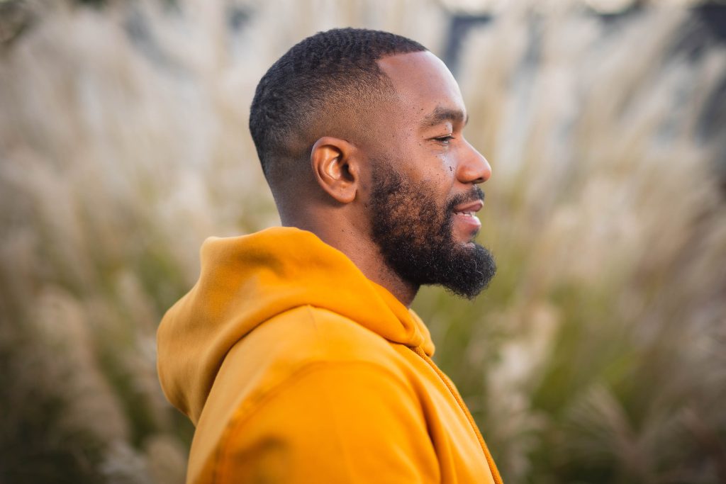 A man in an orange hoodie is standing outdoors, facing to the right. The background is blurry with tall grass, giving the scene a serene portrait-like quality perfect for capturing cherished family moments.