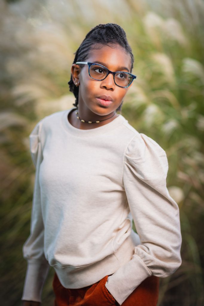 A person wearing glasses and a light-colored sweater stands outdoors with one hand in their pocket, gazing off to the side. The background features tall grass, capturing a serene family portrait moment.