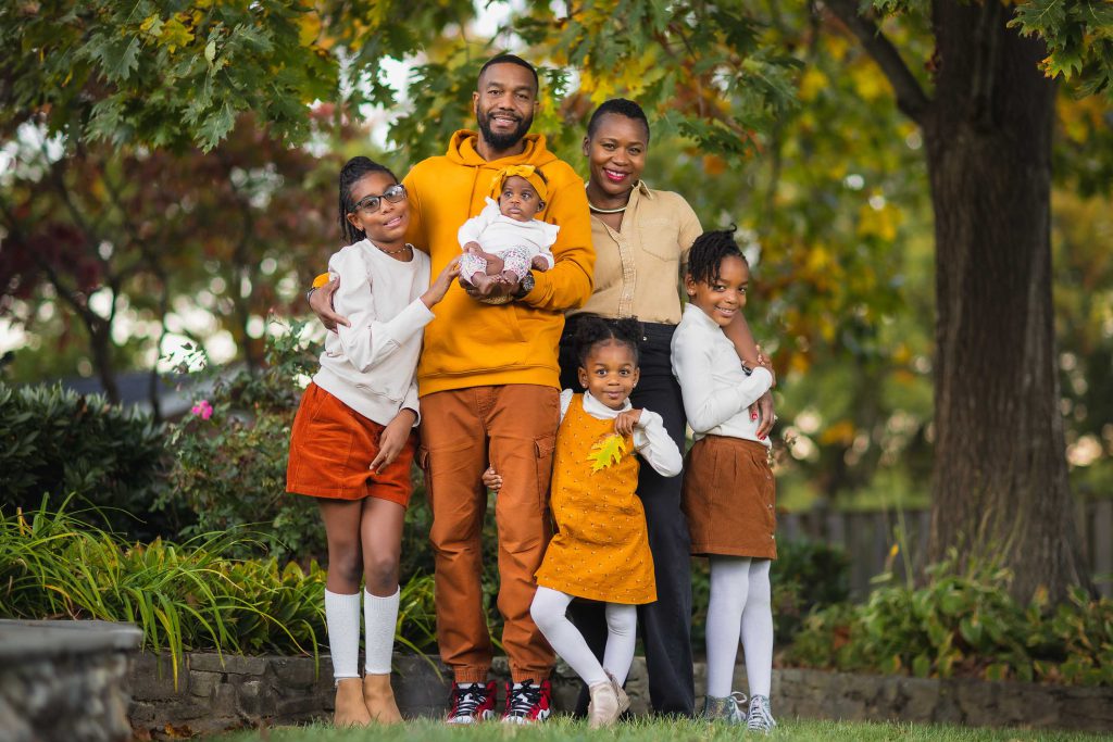 A family of five poses outdoors in autumn for a portrait. The group includes two adults, three children, and an infant. They stand closely together, surrounded by trees and greenery, dressed in coordinated fall attire.