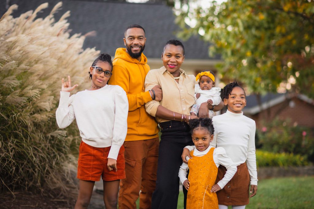A family of six standing outdoors, posing for a portrait. The group includes two adults and four children, all dressed in autumn-toned clothing with a background of grass and trees.
