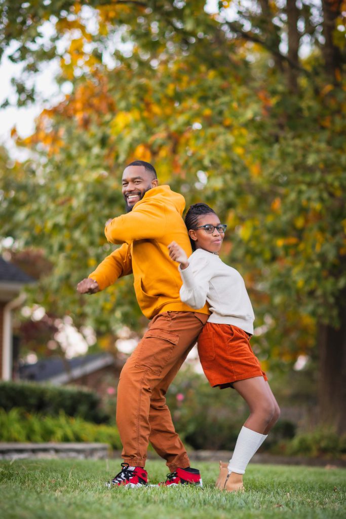 A family portrait features an adult and a child wearing fall-colored clothing, standing back-to-back on a lawn. They smile and pose playfully with autumn trees in the background.