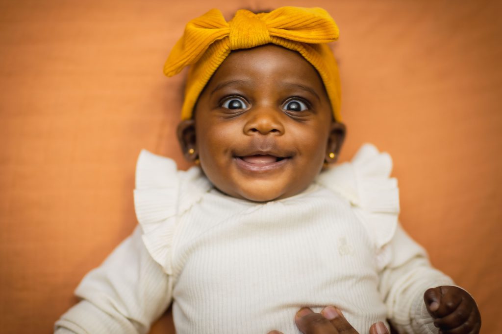 A baby wearing a yellow headband and a white outfit smiles while lying on an orange surface, creating a heartwarming family portrait. A person's hand is visible in the lower right corner.