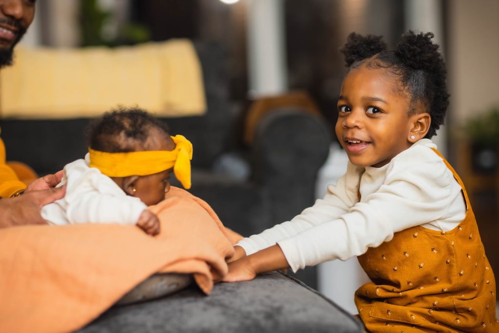 In a charming family portrait, a young child in a white sweater and mustard-colored overalls smiles at the camera while sitting beside a baby lying on a blanket. The baby wears a yellow headband with a bow, completing this heartwarming scene.