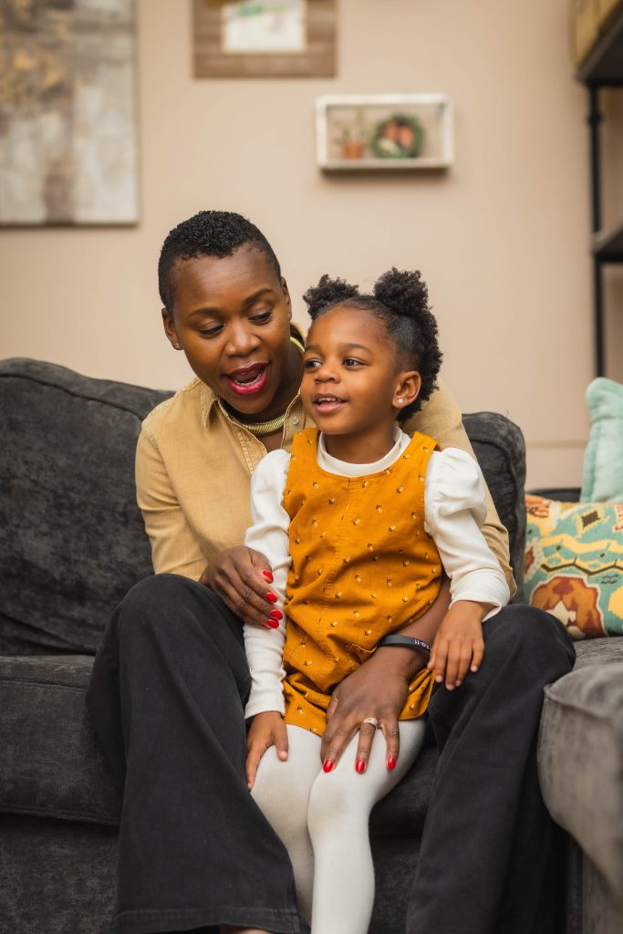 In this heartwarming family portrait, an adult and a child sit together on a couch; the adult holds the smiling child, who is wearing an orange dress over a long-sleeve shirt.