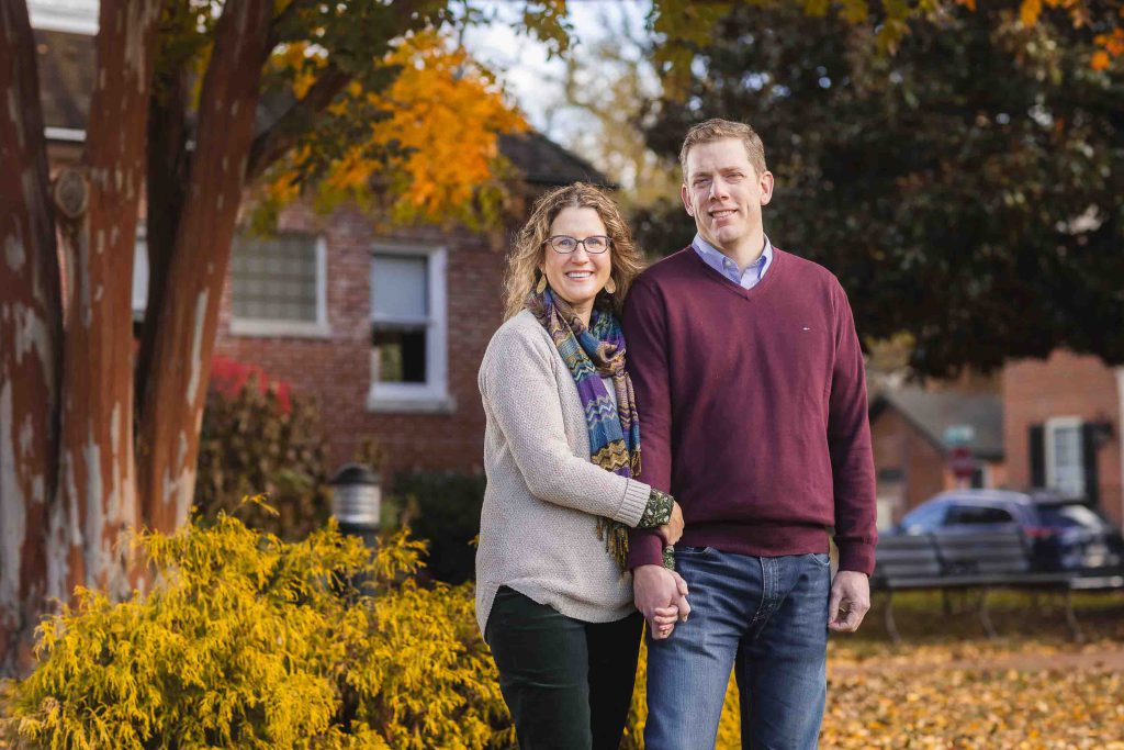 A middle-aged couple, smiling and holding hands, stands in front of autumn foliage in a suburban area for their family portraits.