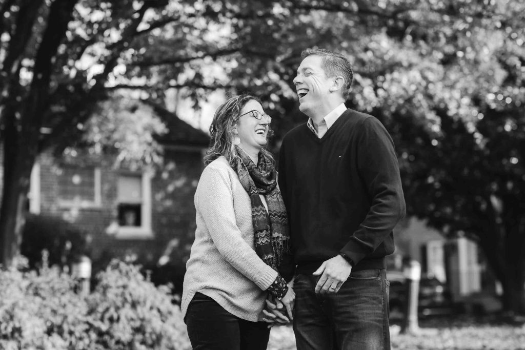A joyful middle-aged couple laughing together in a park, holding hands with trees in the background, in black and white, captured during a family portrait session at Kentlands Mansion.