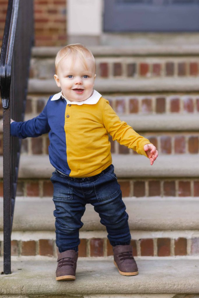Toddler in a color-blocked shirt and jeans standing on steps, holding a railing, with a cheerful expression for family portraits.