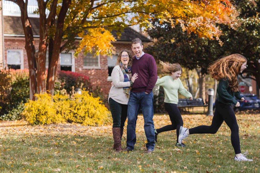 A family of four laughing and playing tag in a park with colorful autumn leaves in the background, perfect for Family Portraits.