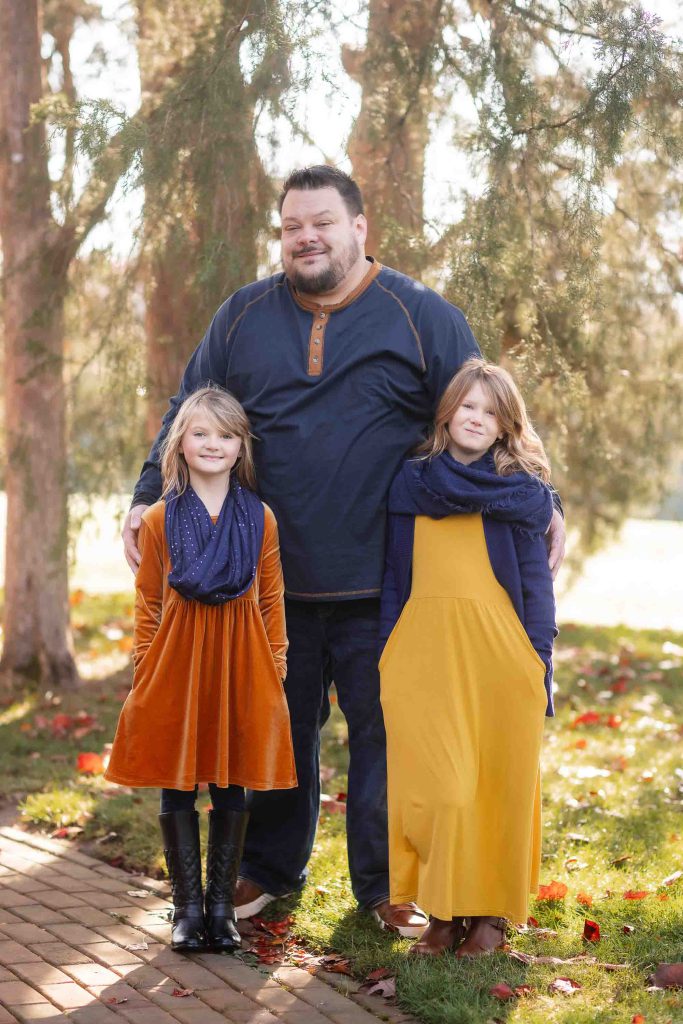 A father stands with his two daughters on a tree-lined path, all three smiling and dressed in autumn colors for their family portraits.