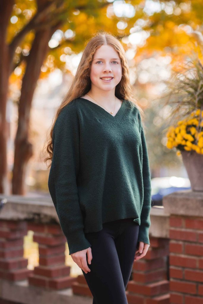A young girl with long curly hair, wearing a green sweater, posing in front of autumn-colored trees for family portraits.