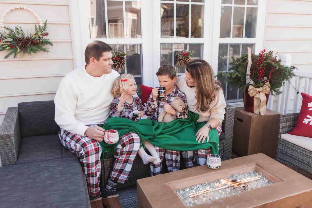 A family of four, dressed in holiday-themed pajamas, enjoying a warm drink together on an outdoor porch decorated for Christmas, perfect for Family Portraits.