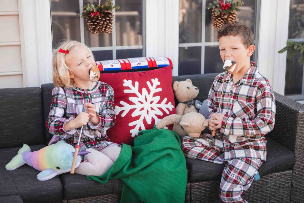 Two children in plaid pajamas sitting on a porch, laughing and eating marshmallows, with a holiday-themed pillow beside them.