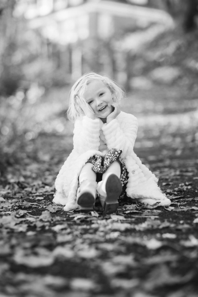 A young girl smiling, sitting on a leaf-covered path, wearing a white coat and sparkly shoes, in a black and white family portrait photo.