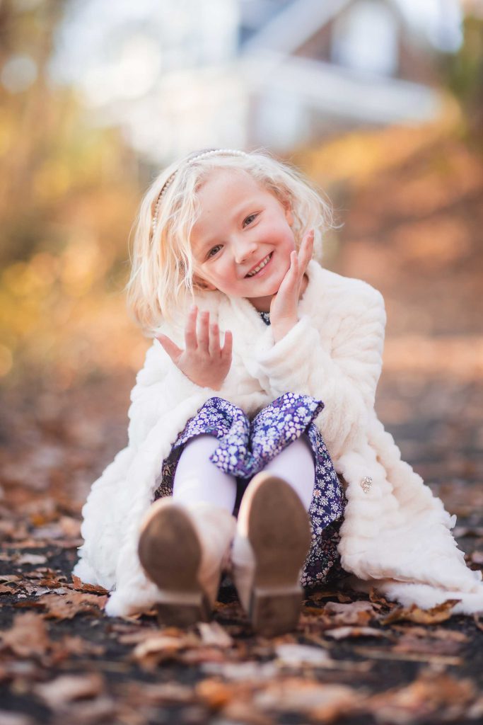 A young girl with blonde hair smiling and waving, sitting on a leaf-covered path in a family portrait, wearing a white coat and patterned dress.