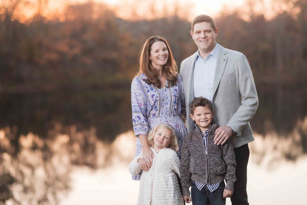 A family of four smiling by a lake at sunset, surrounded by autumn-colored trees, perfect for Family Portraits.