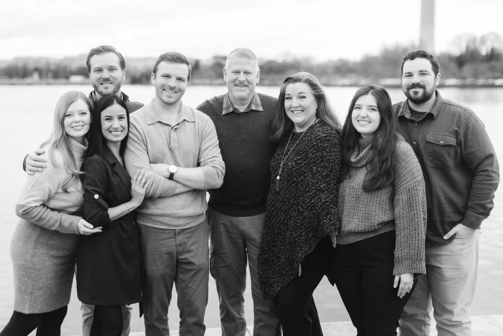 A group of eight people standing in two rows by a body of water, with the Jefferson Memorial towering in the background. Everyone is smiling and wearing casual winter clothing. The black-and-white image has a timeless quality, almost like a family portrait from another era.