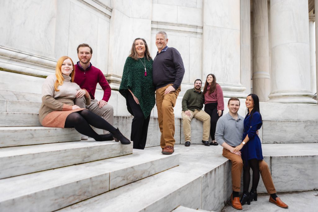 A group of seven people pose on the marble steps of the Jefferson Memorial with large pillars in the background. Four are seated, three are standing, all dressed in casual to semi-formal attire—capturing a timeless family portrait.
