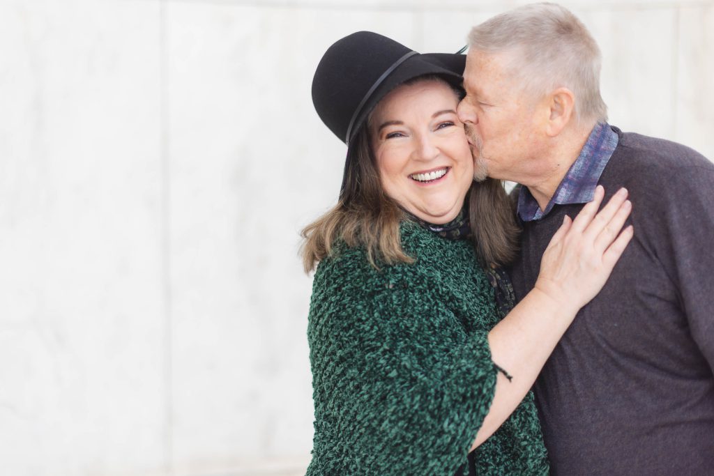 A smiling woman wearing a green shawl and black hat is being kissed on the cheek by an older man in a dark sweater. They stand in front of a light-colored, slightly textured background, evoking the feel of cherished family portraits.