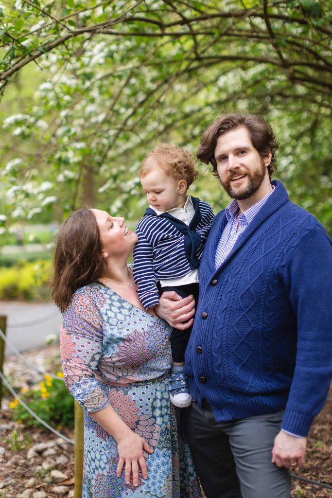 A family of three poses for a portrait under a tree. The man holds a baby as the woman looks up at the child, embodying joy. They are all dressed in blue-toned clothing, capturing an extended moment of connection and warmth.