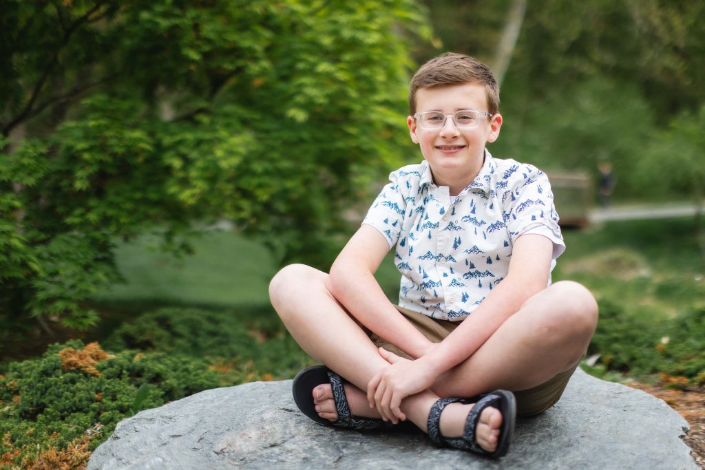 A portrait of a boy with glasses sitting cross-legged on a rock, wearing a white shirt with blue patterns and shorts, outdoors at Brookside Gardens, framed by the lush trees in the background.