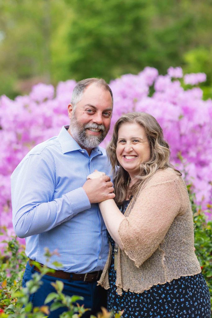 A couple stands outside, smiling and holding hands in front of a blooming garden with pink flowers at Brookside Gardens.