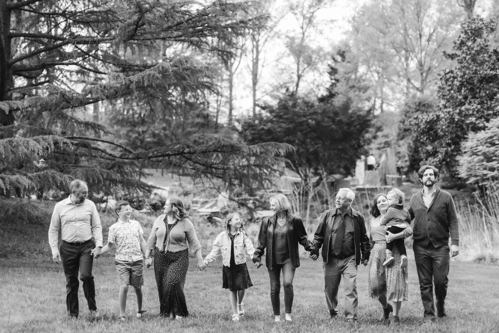 A black-and-white photo captures an extended family of eight holding hands and walking through Brookside Gardens. Four adults walk on either side of four children, with trees visible in the background.