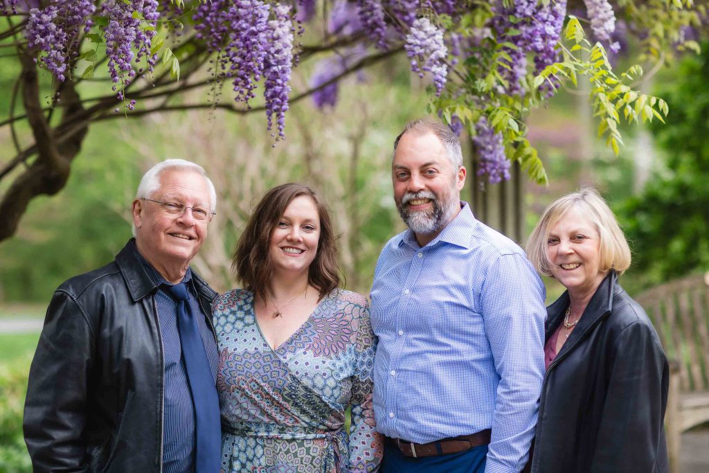 Four people stand smiling under a wisteria tree in what appears to be an extended family portrait. The group includes two men and two women, all dressed in casual to semi-formal attire. A wooden bench is partially visible in the background.