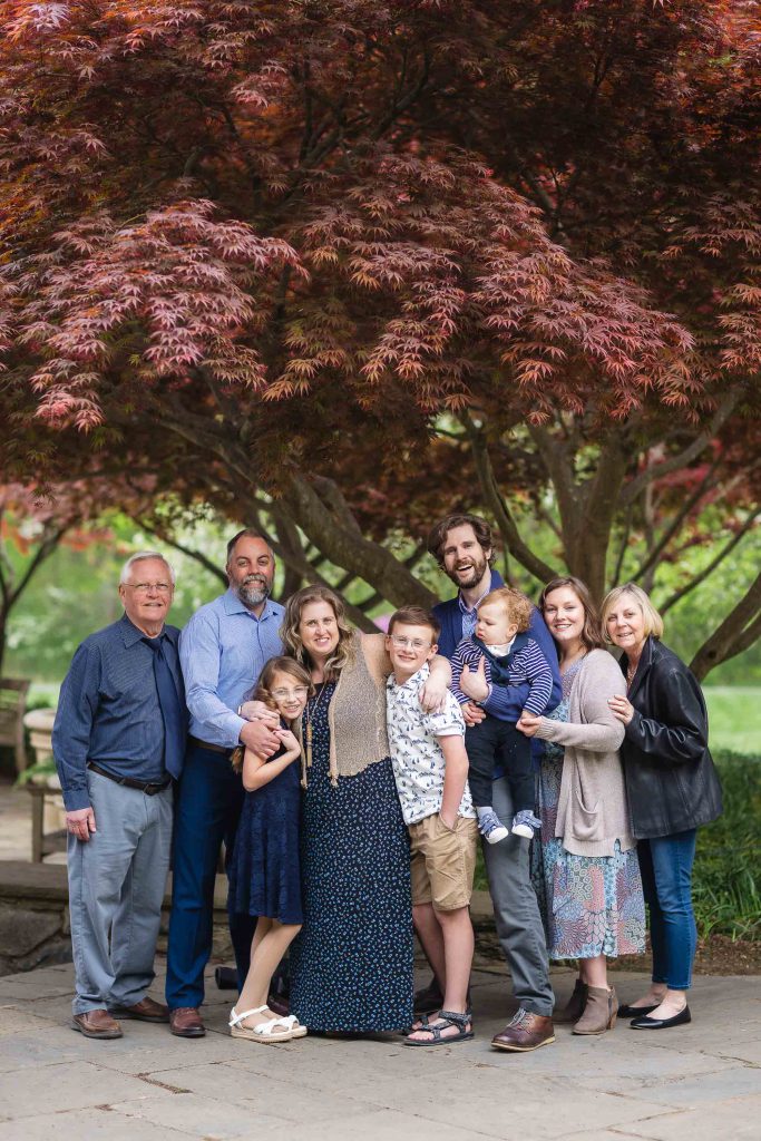 An extended family of nine, including adults and children, pose together under a red-leafed tree at Brookside Gardens.