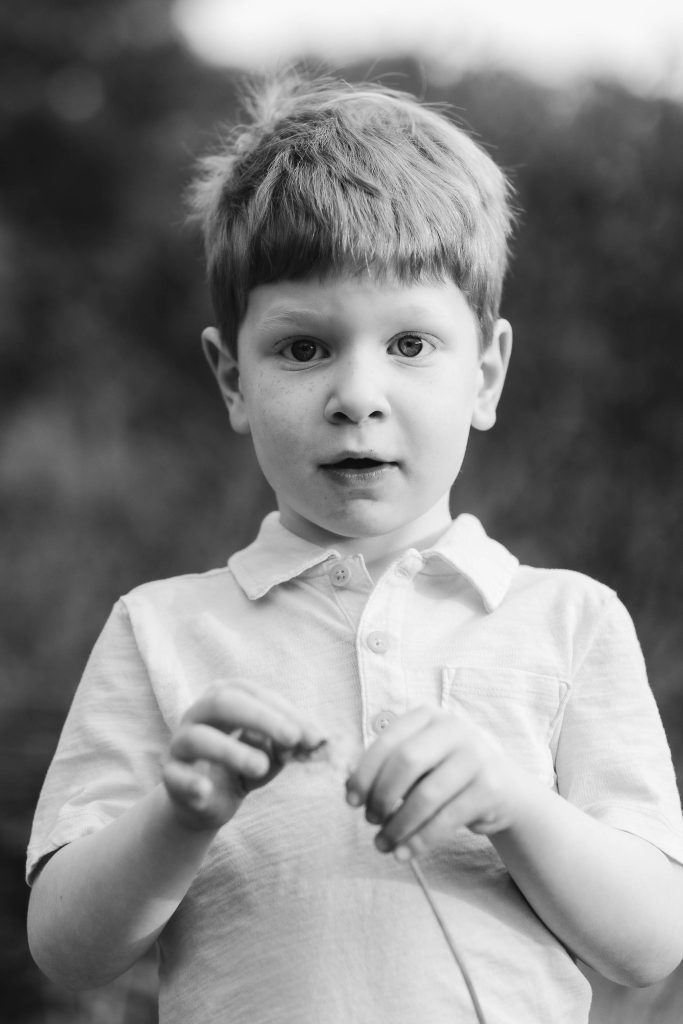 A black and white photo of a young boy with short hair wearing a light-colored shirt. He looks at the camera while holding a small object, capturing a moment of innocence. The blurred background, possibly Quiet Waters Park, adds depth to this timeless family portrait.