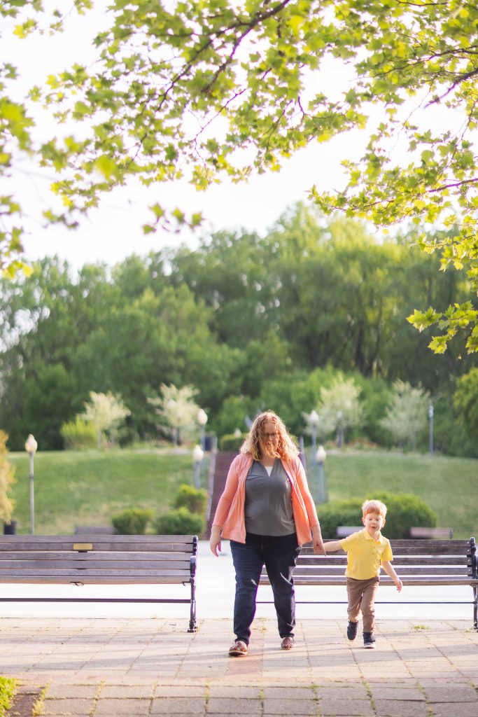 A woman and a young child walk hand-in-hand on a paved path in Quiet Waters Park, with benches on either side and green trees in the background, capturing a serene family portrait.