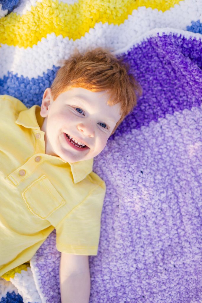 A smiling child with red hair lies on a colorful, striped blanket featuring yellow, white, blue, and purple sections, creating a picture-perfect moment for family portraits.