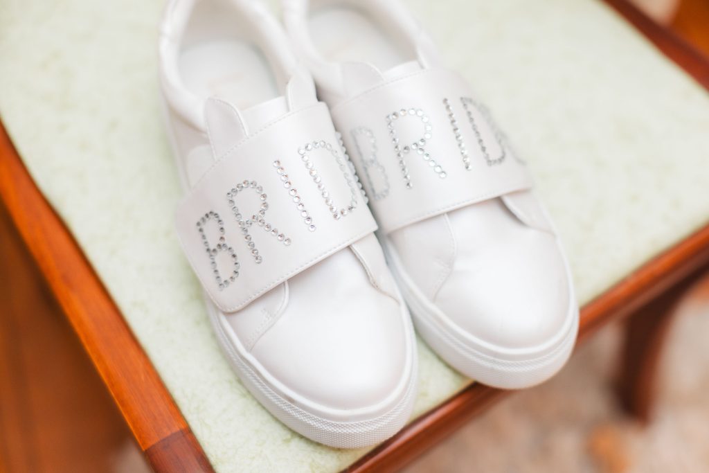 A pair of white sneakers with the word "BRIDE" spelled out in rhinestones on the straps, placed on a wooden chair with a light green cushion, captures the charming details of a wedding at The Barns at Hamilton Station.