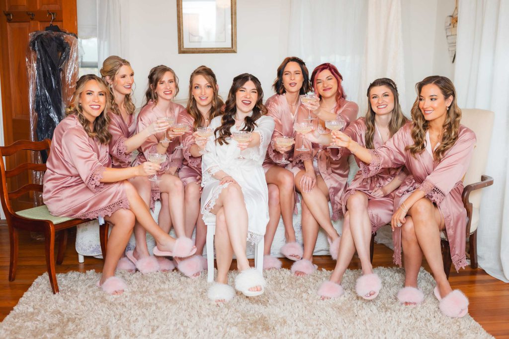 A bride in white sits with eight bridesmaids wearing matching pink robes and slippers, all holding glasses in a well-lit room at The Barns at Hamilton Station. They are seated on a couch and chairs with a rug beneath them, savoring the joyous moments of wedding preparation.