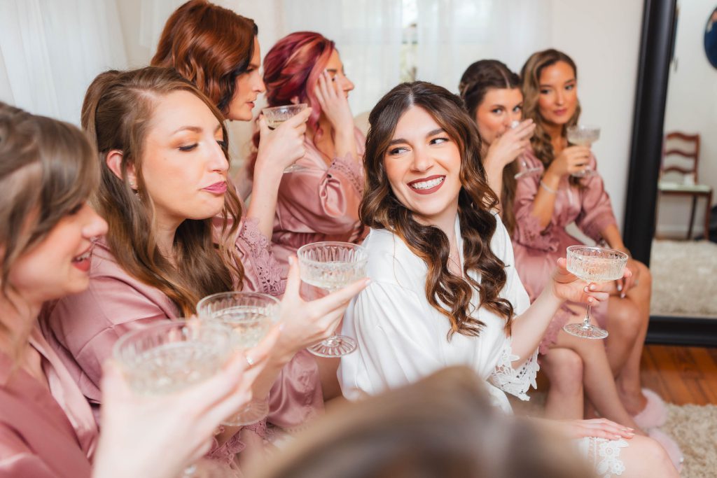 A bride and seven bridesmaids in matching robes smile and raise their glasses in a toast, sitting closely together in a bright room at The Barns at Hamilton Station, excitedly celebrating the wedding preparation.