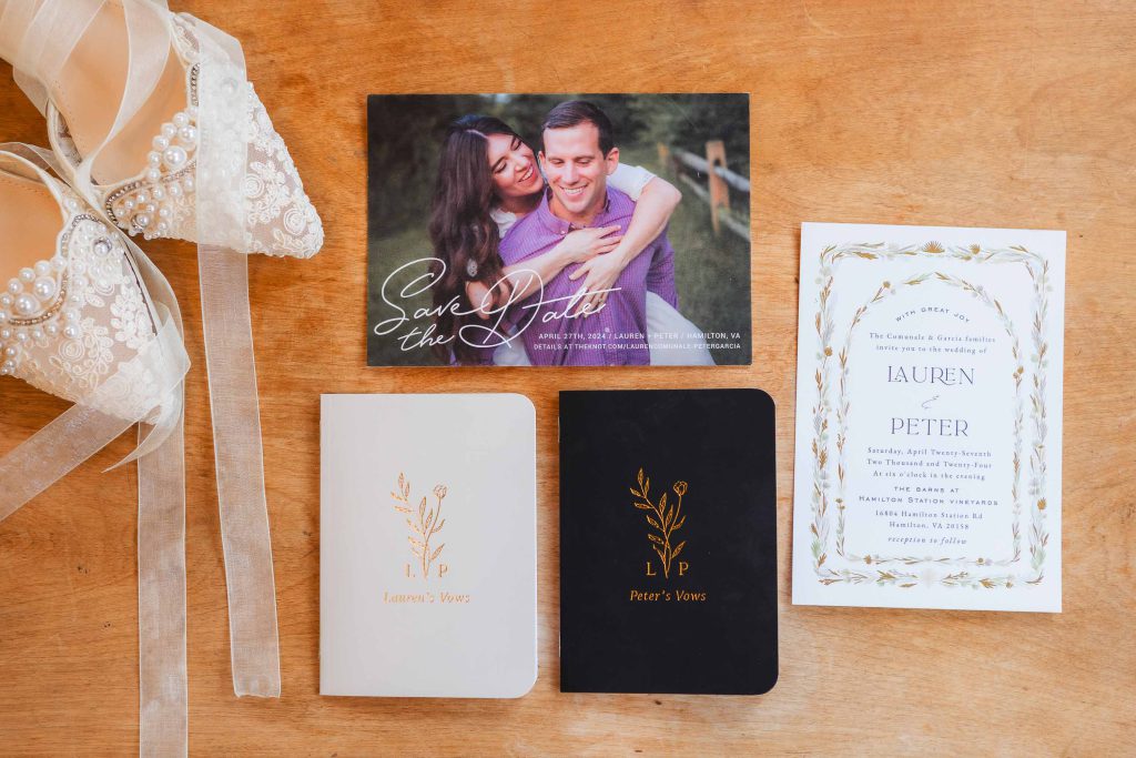 Wedding invitation set displayed on a wooden surface, featuring beige wedding shoes, a "Save the Date" card, an invitation card, and personalized vow booklets for Lauren and Peter. The intricate details hint at their upcoming celebration at The Barns at Hamilton Station.