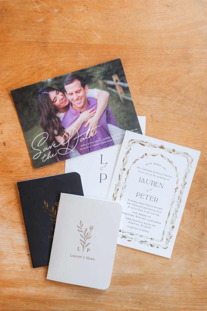 A wooden table at The Barns at Hamilton Station displays a save-the-date card with a couple's photo, a formal wedding invitation, and vow booklets labeled "Lauren's Vows," adorned with botanical designs, showcasing the intricate details of their wedding preparations.