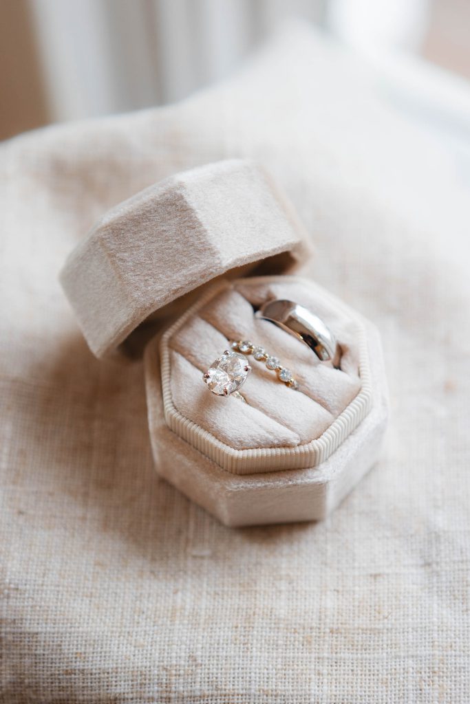 A beige velvet hexagonal ring box, perfect for a wedding at The Barns at Hamilton Station, holds two exquisite rings: one featuring a large oval-shaped gemstone and the other a plain band adorned with small, intricate details.