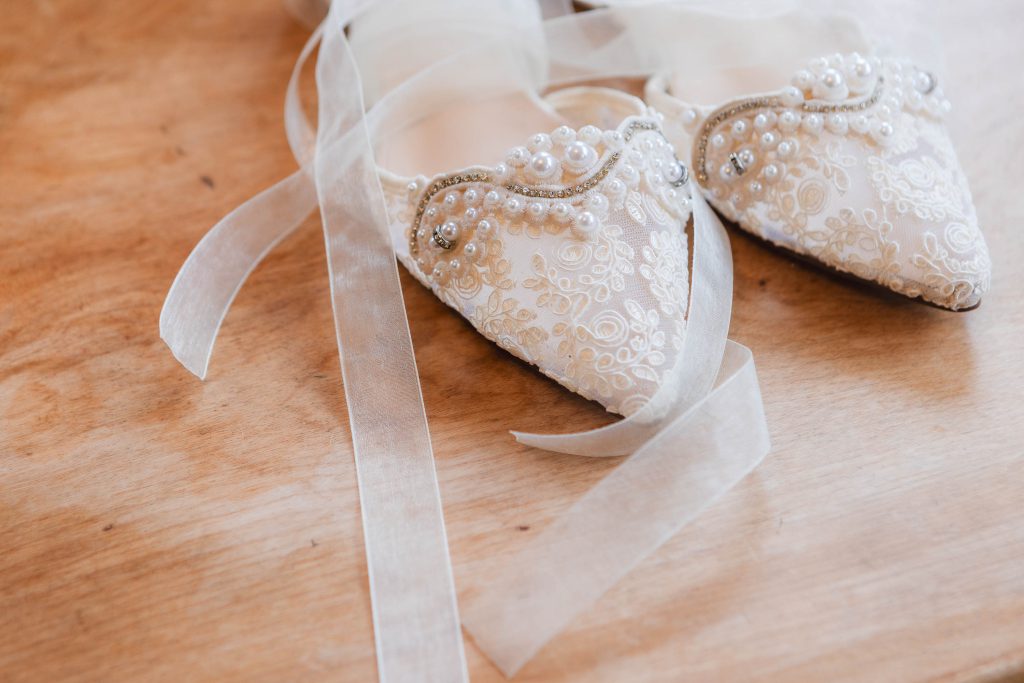Pair of embroidered white shoes with ribbon details, decorated with pearls and beads, placed on a wooden surface at The Barns at Hamilton Station.