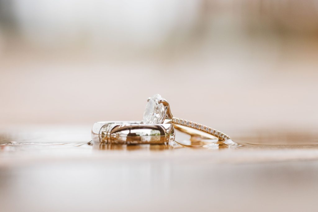 Close-up of two wedding rings, one with a large clear gemstone and a gold band with small inset stones, and the other a plain silver band, placed on a wet wooden surface at The Barns at Hamilton Station, showcasing exquisite details.