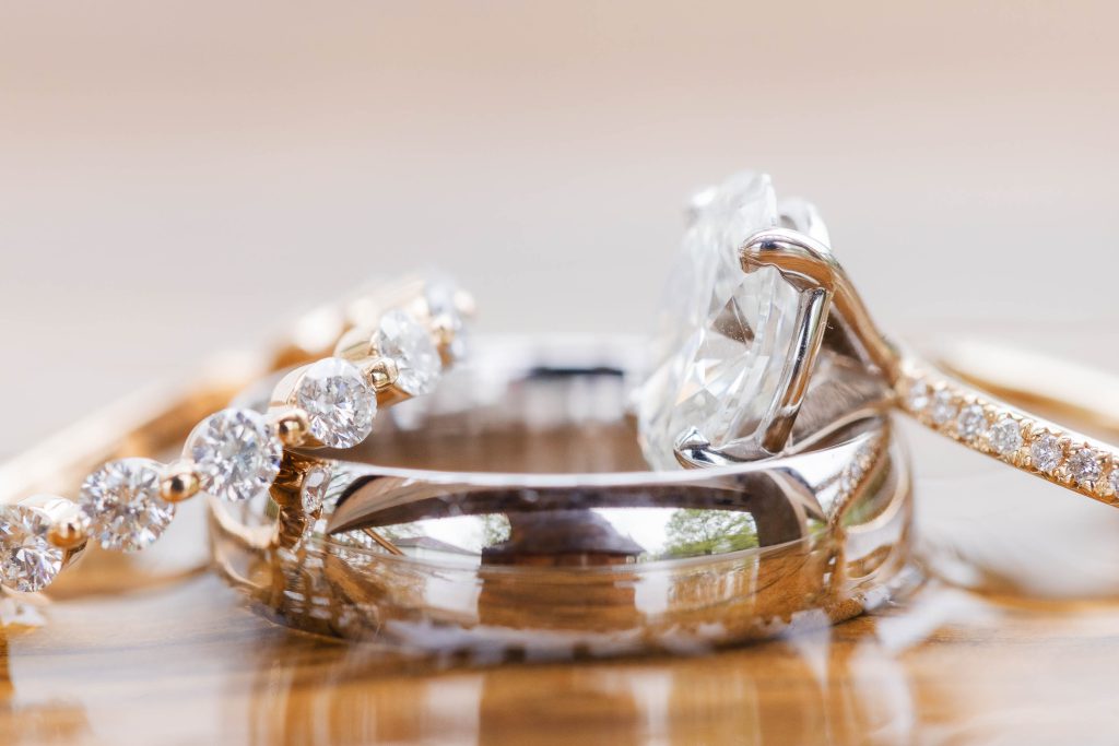 A close-up photo captures the intricate details of three wedding rings on a wooden surface at The Barns at Hamilton Station. Two rings showcase stunning diamond settings, while one is a simple, elegant band.