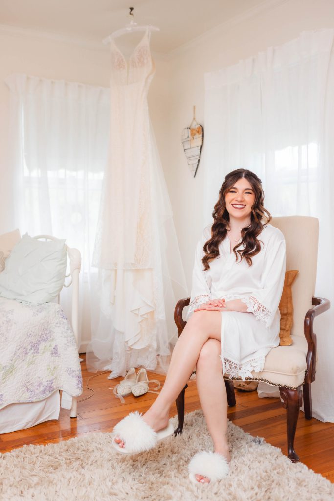 A woman in a white robe and fluffy slippers sits on a chair in a sunlit room at The Barns at Hamilton Station. Behind her, a lace wedding dress hangs gracefully, signaling the final moments of preparation for her special day.