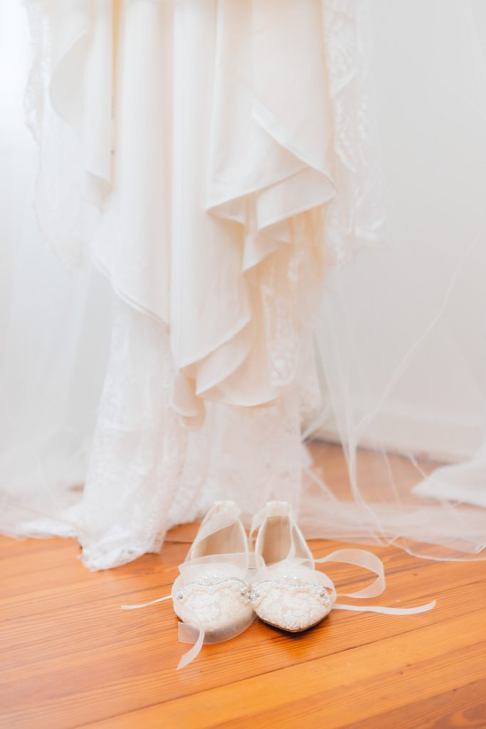 A white dress with lace detailing hangs above a pair of white ballet flats adorned with embellishments, placed on a wooden floor at The Barns at Hamilton Station—capturing the perfect wedding details.
