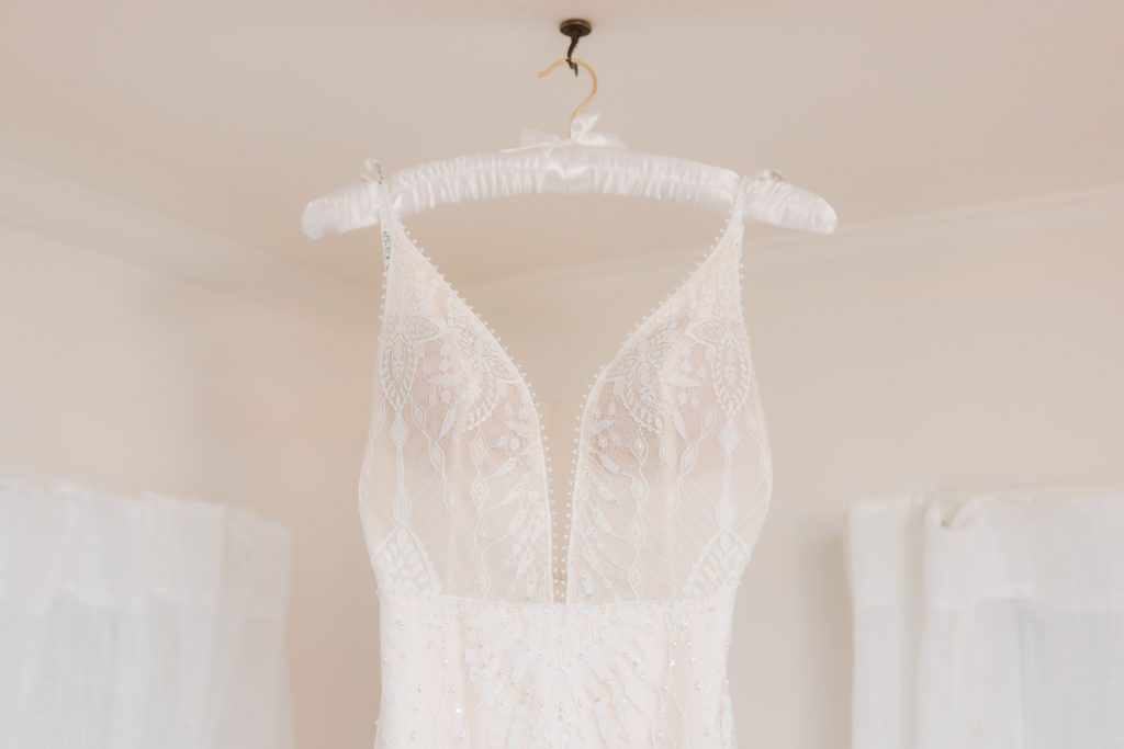 A white lace wedding dress hangs on a white padded hanger against a light-colored backdrop, capturing the intricate details perfect for a wedding at The Barns at Hamilton Station.