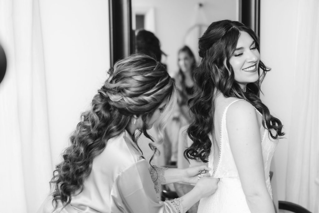 At The Barns at Hamilton Station, a woman in a wedding dress is smiling as another woman helps her with the back of the dress. They are standing in front of a mirror, immersed in the joyous preparation for the wedding day.
