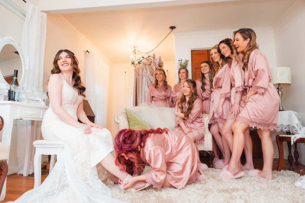 In a bright, cozy room at The Barns at Hamilton Station, bridesmaids in pink robes surround a smiling bride in a white gown. The joyous wedding preparation unfolds as one bridesmaid helps the bride with her shoe.