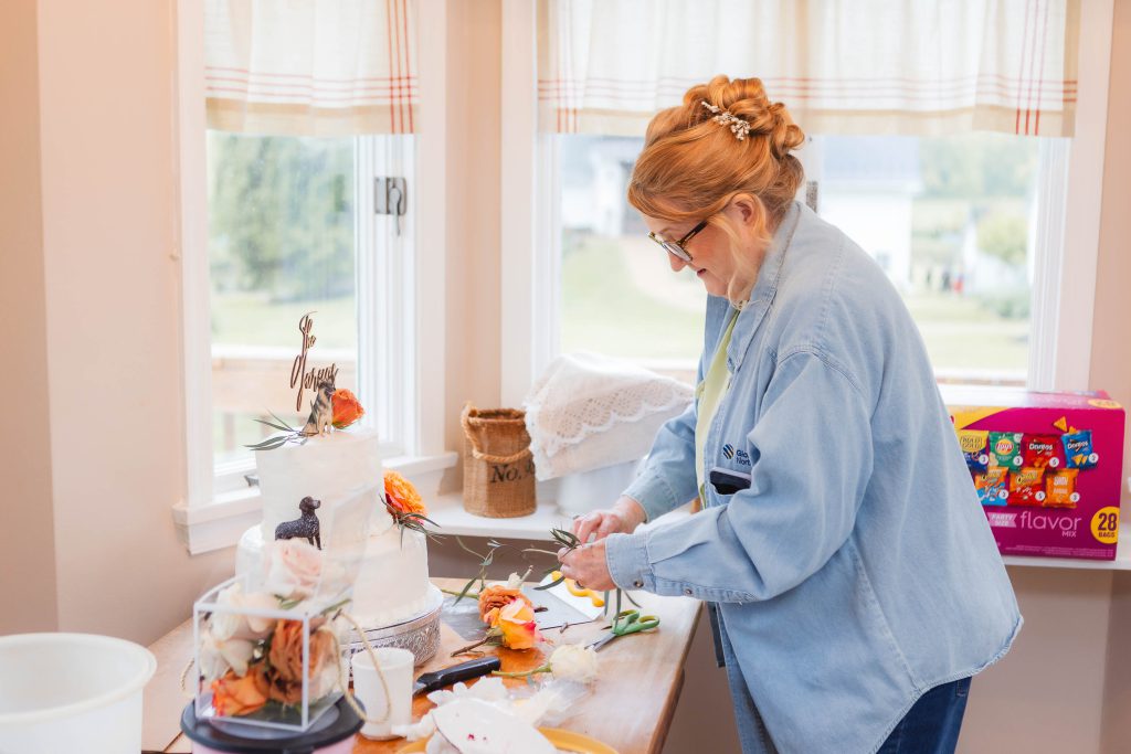 A person trims flowers and decorates a two-tiered white wedding cake on a table near a window. The scene, set at The Barns at Hamilton Station, includes cake decorating tools, floral decorations, and a box of flavor packets in the background.